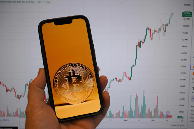 This illustration photograph shows a Bitcoin cryptocurrency coin picture displayed on a smartphone beside a screen showing a trading chart in Brussels on Tuesday (AFP photo)