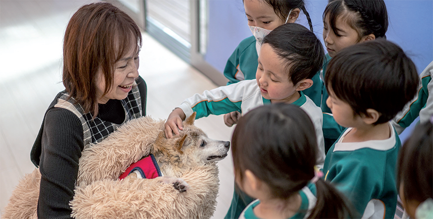 This photo taken on March 19 shows Atsuko Sato (left) with her Japanese shiba inu dog Kabosu, best known as the logo of cryptocurrency Dogecoin, playing with students at a kindergarten in Narita, Chiba prefecture, east of Tokyo (AFP photo)