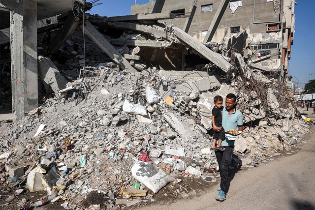 A man carries a child while walking past a collapsed building in the Bureij camp for Palestinian refugees in the central Gaza Strip on October 9, 2024 (AFP photo)