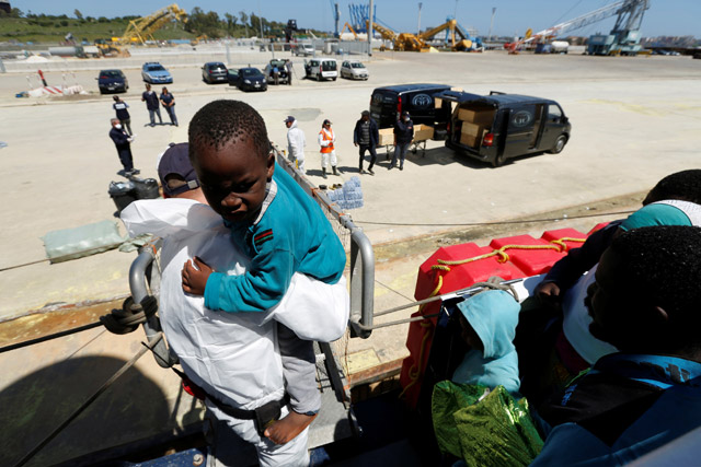 A crewman of the Malta-based NGO Migrant Offshore Aid Station ship Phoenix helps a migrant child to disembark as coffins with the bodies of dead migrants are put in a hearse, after the ship arrived with migrants and bodies on board in Augusta, Italy, on Wednesday (Reuters photo)