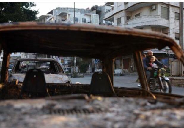 Members of a family riding a motorcycle drive past a charred car in the town of Jableh in Syria's coastal province of Latakia on March 12, 2025 (AFP photo)