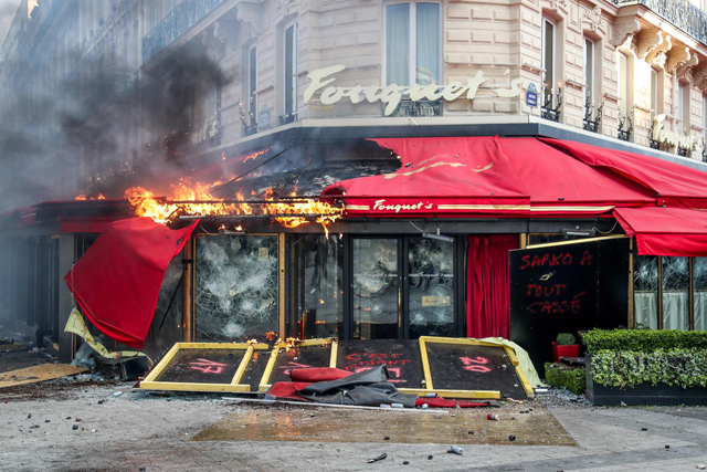 In this file photo taken on March 16, people stand outside the restaurant ‘Le Fouquet's’ set on fire during clashes with riot police forces on the Champs-Elysees in Paris (AFP photo)