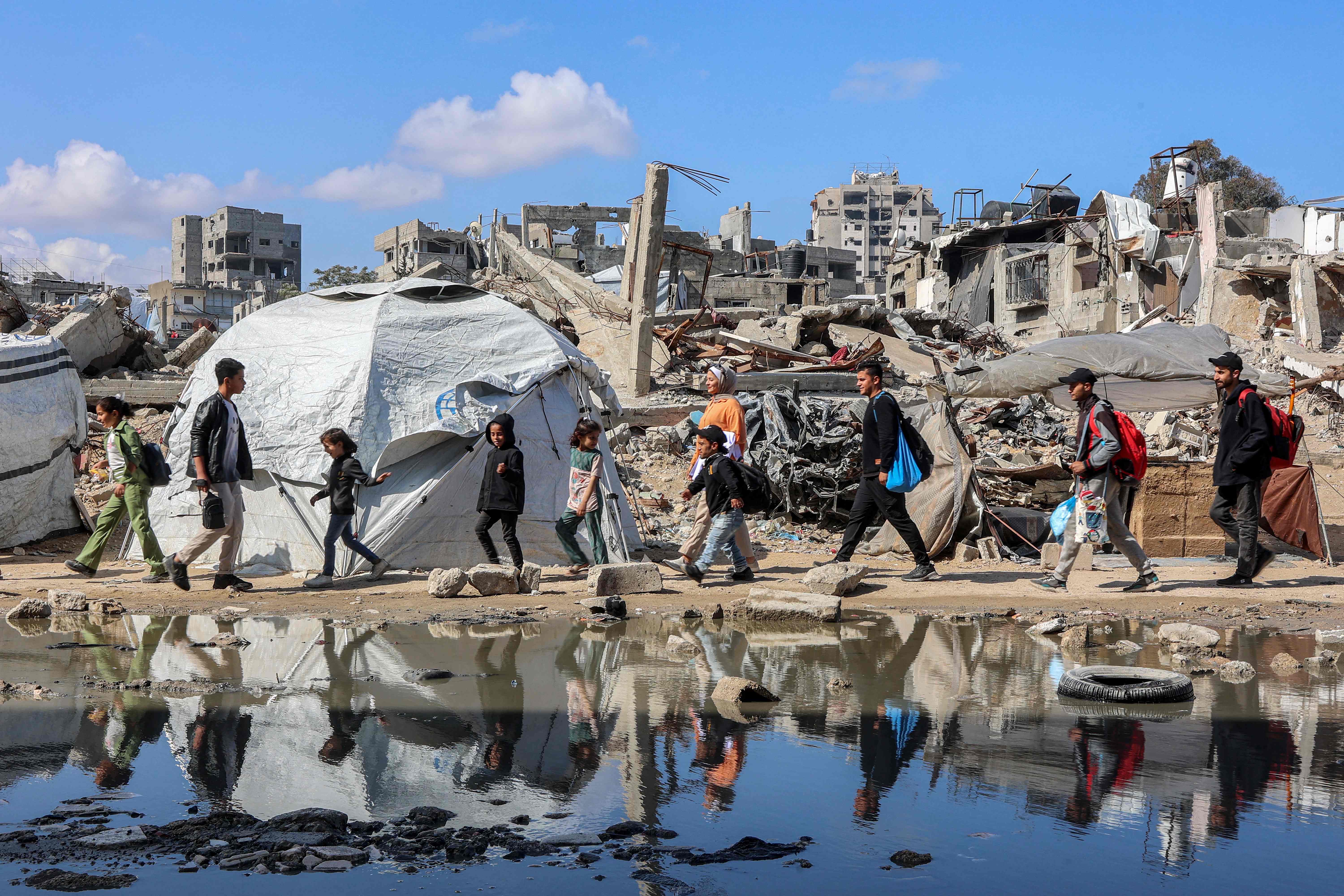 People walk past a puddle of water by a tent shelter erected near the rubble of a collapsed building in the Nasr neighbourhood in western Gaza City on April 15, 2025 (AFP photo)