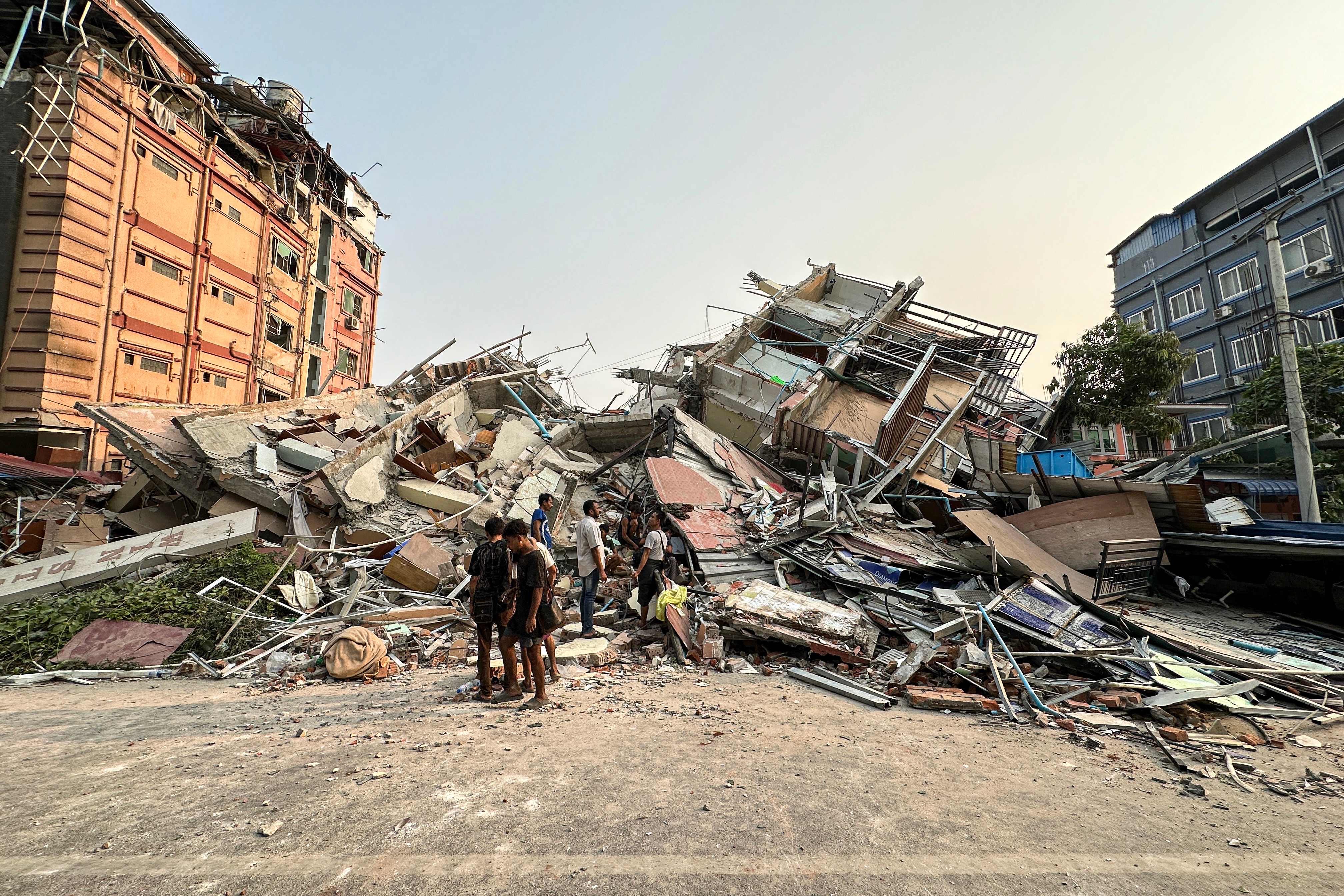 People stand past the debris of a collapsed building in Mandalay on March 28, 2025, after an earthquake