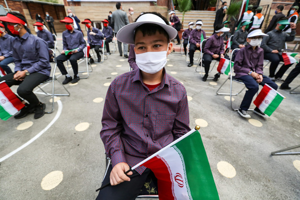 Mask-clad school children hold Iranian national flags as they sit distanced apart from each other in the yard of a school while attending a speech by Iran's president by video conference, at Nojavan school in the capital Tehran on the first day of schools reopening, on Saturday (AFP photo)