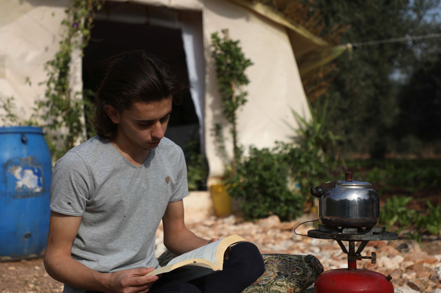 Displaced Syrian, Wissam Diab, 19, reads a book sited in front of his new home, a tent surrounded by luscious plants, which recreates his childhood home, in the town of Atme in Syria’s north-western Idlib province, on Thursday (AFP photo)