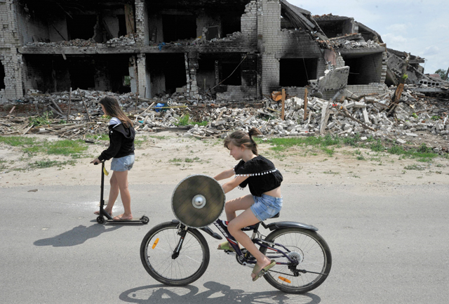 Children ride a bike and a scooter on a road in front of a destroyed building in the village of Novoselivka, outside Chernigiv, on Tuesday, amid the Russian invasion of Ukraine (AFP photo)