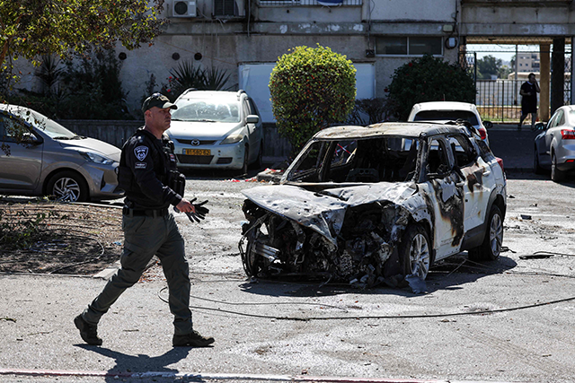 A member of the security forces walks in font of a charred car near a building hit by a rocket fired from Lebanon in Kiryat Ata in Haifa on October 19, 2024 (AFP)